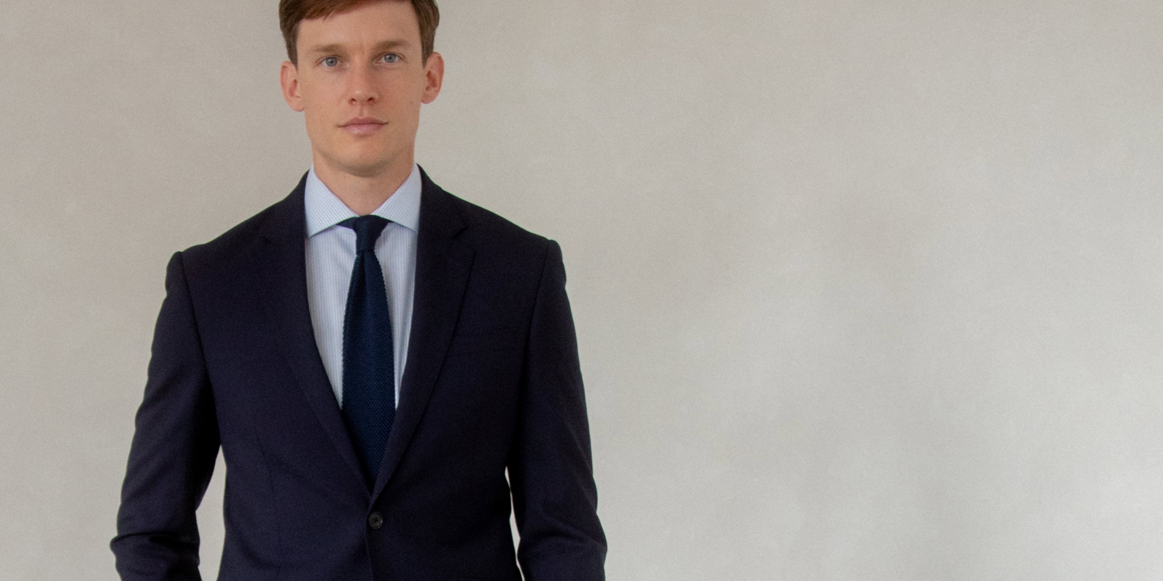 A male model wears a navy blue suit with a blue striped shirt and knitted navy tie.