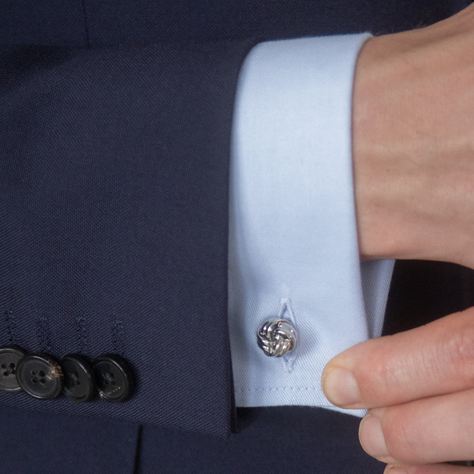 A silver metal knoted cufflink holds together the french double cuff of a light blue twill shirt. The shirt cuff sit  proud of a buttoned navy suit cuff.